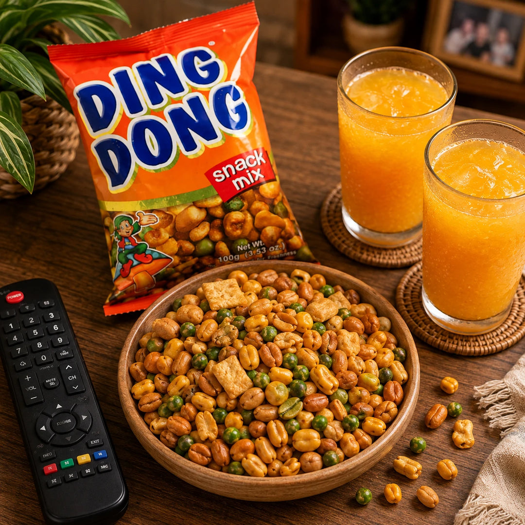 A bowl of Ding Dong snack mix with peanuts, green peas, and crackers, next to an orange bag of Ding Dong snack mix, two glasses of orange juice, and a remote control on a wooden table.