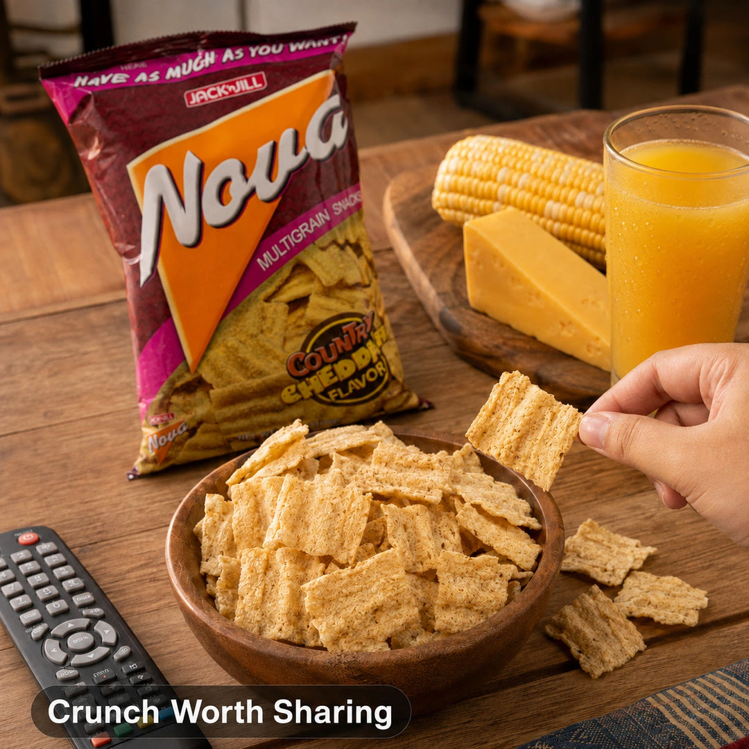 A bag of Jack 'n Jill Nova multigrain cheddar-flavored snack chips stands on a wooden table beside a bowl of chips, a hand holding a chip, corn, cheese, and a glass of orange juice.
