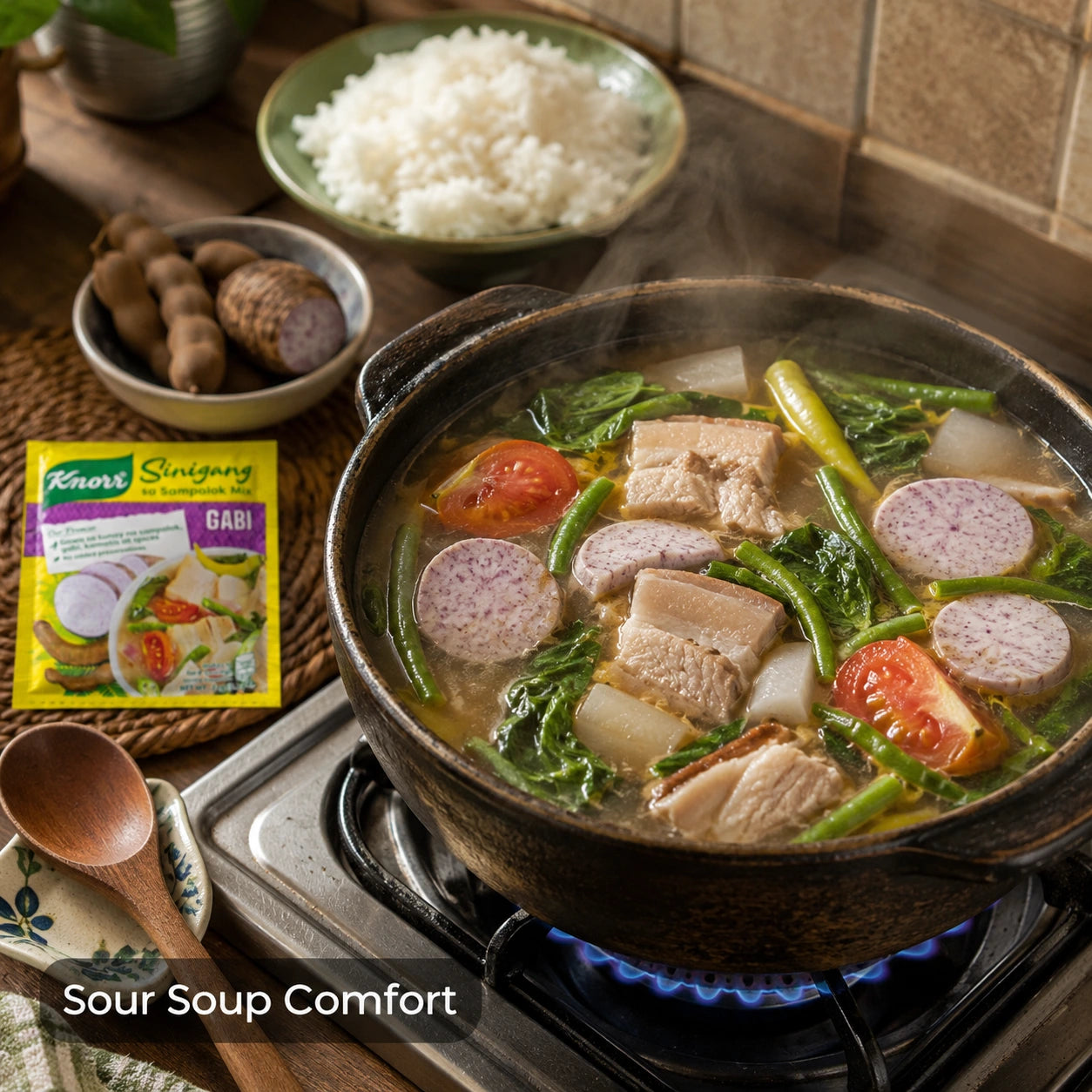 A pot of sour pork sinigang soup comfort hotpot simmering on a gas stove, filled with pork belly, greens, and tomatoes, with a packet of knorr sinigang mix with gabi and bowl of rice nearby on a kitchen counter.