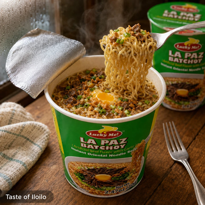 A cup of Lucky Me! La Paz Batchoy instant noodles on a rustic wooden table, with a fork lifting a portion of the noodles that include a raw egg yolk. Another cup is visible in the background, and a small 'Taste of Iloilo' label appears in the bottom-left corner.