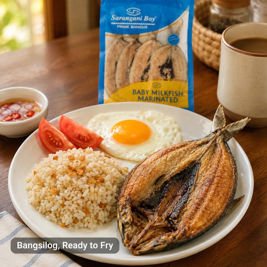 A plated breakfast scene on a wooden table featuring a cooked bangsilog fish piece, rice, fried egg, tomato wedges, a small bowl of sauce, and a package of Sarangani Bay Baby Milkfish Marinated snack in the background.